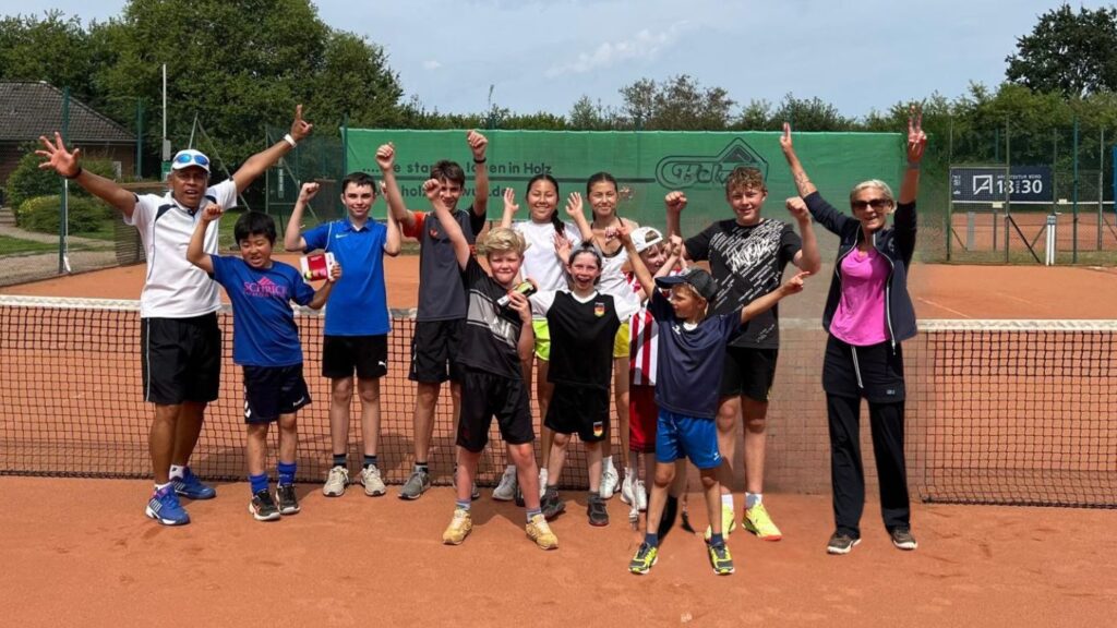 Jubelnde Kinder und zwei Tennis-Trainer:innen auf einem Tennis-Platz